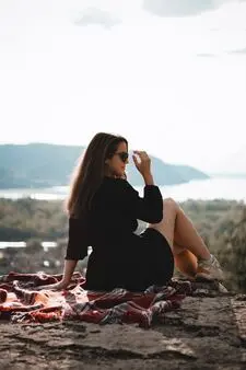 A smiling woman sits on the cliff, wearing a black dress and sunglasses