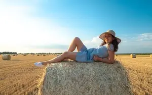 A woman wearing a farm outfit with a hat lies with a smile on a bale of hay