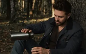 A man is sitting against a tree and pouring liquid from his flask